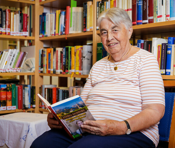 Eine ältere Dame sitzt vor einem Bücherregal, hat ein aufgeschlagenes Buch in der Hand und lächelt in die Kamera.