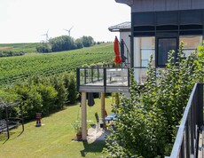 Das Bild zeigt den Balkon und Garten des Hauses Lisa mit Blick auf weitläufige Weinberge und Windräder in der Ferne. Die Umgebung wirkt grün und friedlich.