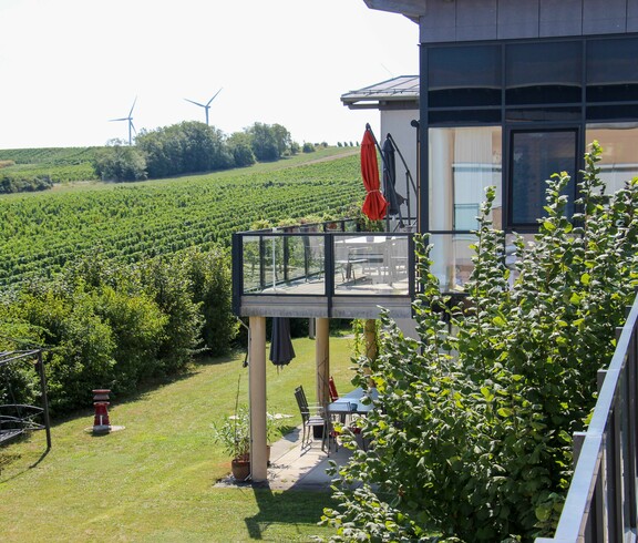 Das Bild zeigt den Balkon und Garten des Hauses Lisa mit Blick auf weitläufige Weinberge und Windräder in der Ferne. Die Umgebung wirkt grün und friedlich.