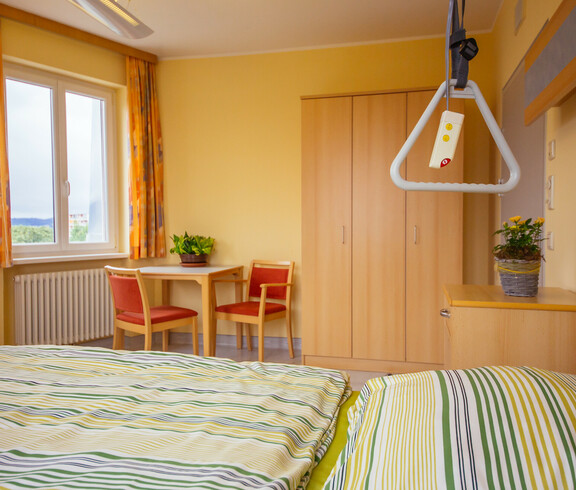 In einem hellen Zimmer stehen ein Tisch und zwei Stühle, Nachtkästchen, Kleiderschrank, Fenster, orange Vorhänge, Heizkörper und ein Bett für pflegebedürftige Personen mit frisch überzogener gelb-grün gestreifter Bettwäsche. Auf dem Tisch und dem Nachtkästchen befindet sich jeweils eine Zimmerpflanze. 