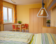 In einem hellen Zimmer stehen ein Tisch und zwei Stühle, Nachtkästchen, Kleiderschrank, Fenster, orange Vorhänge, Heizkörper und ein Bett für pflegebedürftige Personen mit frisch überzogener gelb-grün gestreifter Bettwäsche. Auf dem Tisch und dem Nachtkästchen befindet sich jeweils eine Zimmerpflanze. 