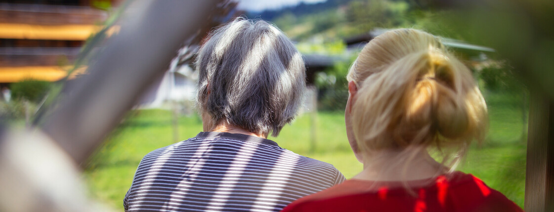 Eine ältere Dame und eine junge Frau sitzen nebeneinander in einer Laube, beide sieht man nur von hinten.