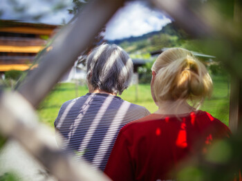 Eine ältere Dame und eine junge Frau sitzen nebeneinander in einer Laube, beide sieht man nur von hinten.