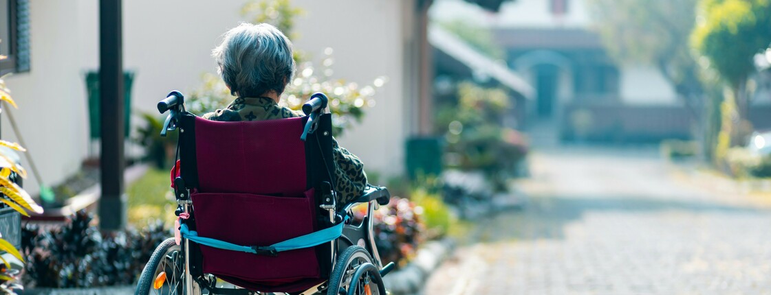 Eine im Rollstuhl sitzende Seniorin befindet sich in einer Hauseinfahrt und schaut die Straße hinunter.