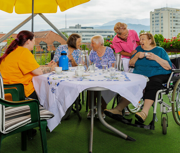 Eine Gruppe von Bewohnerinnen sitzt gemeinsam mit zwei Pflegerinnen bei einer Tasse Kaffee auf der Dachterrasse des Haues. 