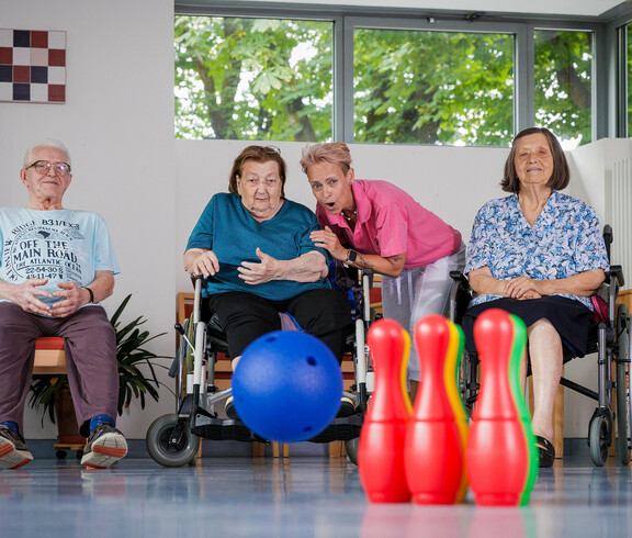 Eine Gruppe von Bewohner*innen sitzt gemeinsam vor Pins eines Bowlingspieles, eine Bewohnerin hat soeben den Ball geworfen, der Richtung Pins rollt. Eine Betreuerin feuert die Gruppe beim Spiel an.