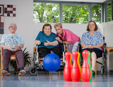 Eine Gruppe von Bewohner*innen sitzt gemeinsam vor Pins eines Bowlingspieles, eine Bewohnerin hat soeben den Ball geworfen, der Richtung Pins rollt. Eine Betreuerin feuert die Gruppe beim Spiel an.