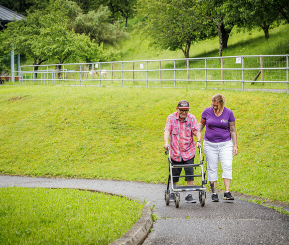 Eine Herr mit Rollator geht mir einer Betreuerin auf dem asphaltierten Weg spazieren.