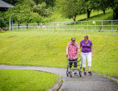 Eine Herr mit Rollator geht mir einer Betreuerin auf dem asphaltierten Weg spazieren.