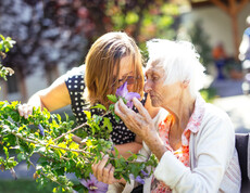 Eine junge Frau und eine ältere Dame riechen an einer Blume des Eibisch-Strauches im Garten.