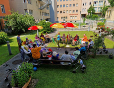 Eine Gruppe von Bewohner*innen sitzt in einem Kreis im Garten und mimt die Bewegungen mit den Tüchern der Betreuerin in der Mitte nach.