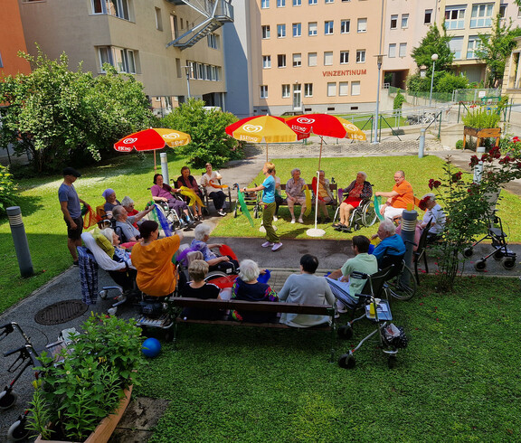 Eine Gruppe von Bewohner*innen sitzt in einem Kreis im Garten und mimt die Bewegungen mit den Tüchern der Betreuerin in der Mitte nach.