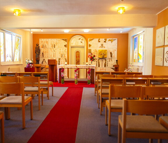 Ein langer roter Teppich führt zu einer schönen Kapelle. Es befindet sich ein Altar mit Kerzen, Blumen, Figur der Heiligen Maria mit Jesuskind, Gemälden, bunten Kirchenfenstern und ausreichend Sitzgelegenheiten.