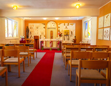 Ein langer roter Teppich führt zu einer schönen Kapelle. Es befindet sich ein Altar mit Kerzen, Blumen, Figur der Heiligen Maria mit Jesuskind, Gemälden, bunten Kirchenfenstern und ausreichend Sitzgelegenheiten.