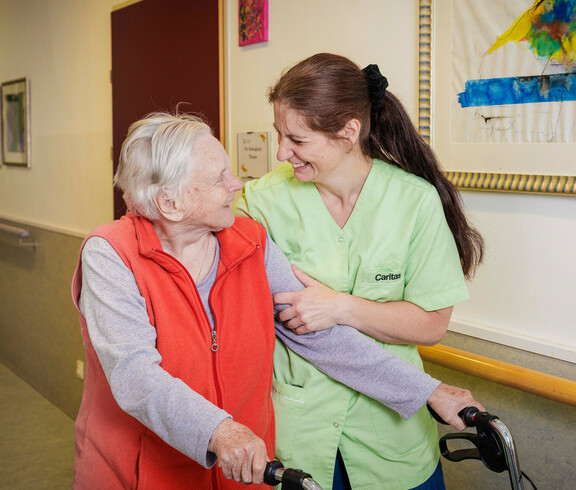 Eine Pflegerin stützt eine Bewohnerin während diese mit dem Rollator den Gang des Pflegewohnhauses entlang geht.