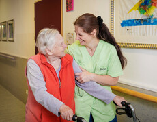 Eine Pflegerin stützt eine Bewohnerin während diese mit dem Rollator den Gang des Pflegewohnhauses entlang geht.