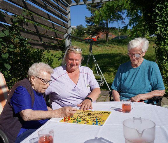 Zwei Bewohnerinnen sitzen zusammen mit einer Pflegekraft draußen an einem Tisch und spielen ein Brettspiel. Alle drei wirken konzentriert und genießen die Aktivität im Freien, während sie bei sonnigem Wetter beisammen sind.