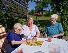 Zwei Bewohnerinnen sitzen zusammen mit einer Pflegekraft draußen an einem Tisch und spielen ein Brettspiel. Alle drei wirken konzentriert und genießen die Aktivität im Freien, während sie bei sonnigem Wetter beisammen sind.