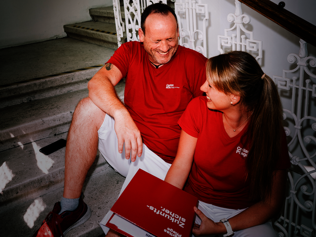 Ein Mann und eine Frau mit roten T-Shirts mit Caritas-Logo sitzen auf den Stiegen und lachen gemeinsam. Die Frau hat einen Aktenordner in der Hand.