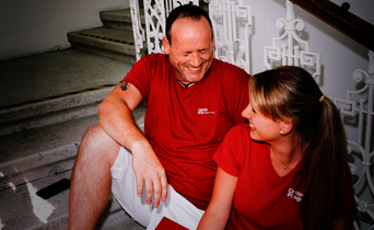 Ein Mann und eine Frau mit roten T-Shirts mit Caritas-Logo sitzen auf den Stiegen und lachen gemeinsam. Die Frau hat einen Aktenordner in der Hand.