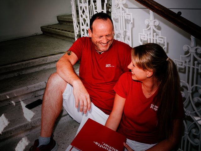 Ein Mann und eine Frau mit roten T-Shirts mit Caritas-Logo sitzen auf den Stiegen und lachen gemeinsam. Die Frau hat einen Aktenordner in der Hand.