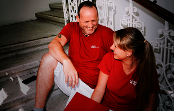 Ein Mann und eine Frau mit roten T-Shirts mit Caritas-Logo sitzen auf den Stiegen und lachen gemeinsam. Die Frau hat einen Aktenordner in der Hand.