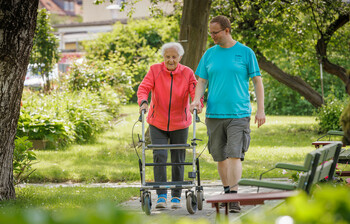 Eine ältere Dame begleitet von einem Pfleger gehen im Garten spazieren.