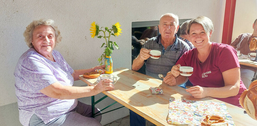 Besucher*innen des Erntedankfest im Haus Elisabeth sitzen gemeinsam an einem Holztisch im Freien und trinken Kaffee. Sie wirken entspannt und genießen die Gesellschaft. Auf dem Tisch stehen Sonnenblumen in einer Vase und einige Brezeln. Alle drei Personen lächeln freundlich in die Kamera.