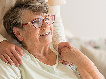 Eine Seniorin mit Brille sitzt in einem Rollstuhl und hält die Hand einer Angehörigen, welche hinter ihr steht.