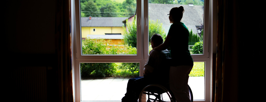 Eine Frau steht mit einem Menschen im Rollstuhl, die nur als Schatten gesehen werden, weil sie vor einem beleuchtenden Fenster stehen.