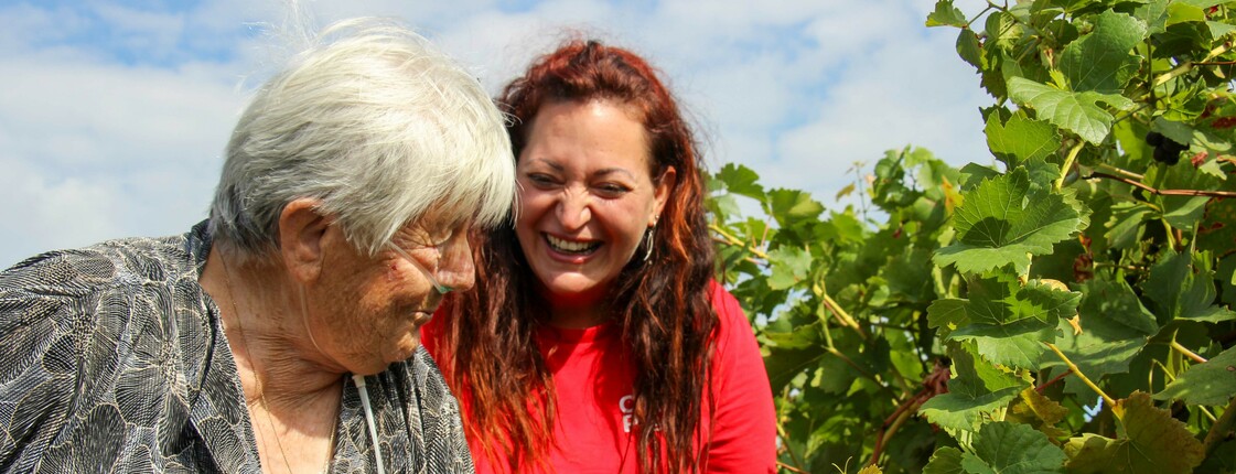 Eine Bewohnerin vom Haus Lisa und eine Betreuerin im roten Caritas Shirt stehen lachend in einem Garten neben Weinreben. Die Stimmung ist herzlich und fröhlich, und die beiden scheinen den Moment in der Natur zu genießen.