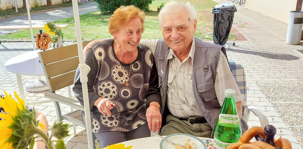 Zwei Personen genießen das Erntedankfest im Haus Elisabeth, indem sie gemeinsam an einen Tisch im Freien sitzen und in die Kamera lächeln. Vor ihnen stehen eine Tasse Kaffee, ein Stück Kuchen und eine Flasche Wasser. 