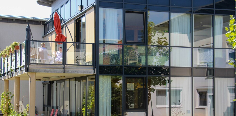 Außenansicht des Hauses Lisa mit großen Glasfronten und einem Balkon, auf dem zwei Personen sitzen. Zu sehen ist auch eine Grünfläche.