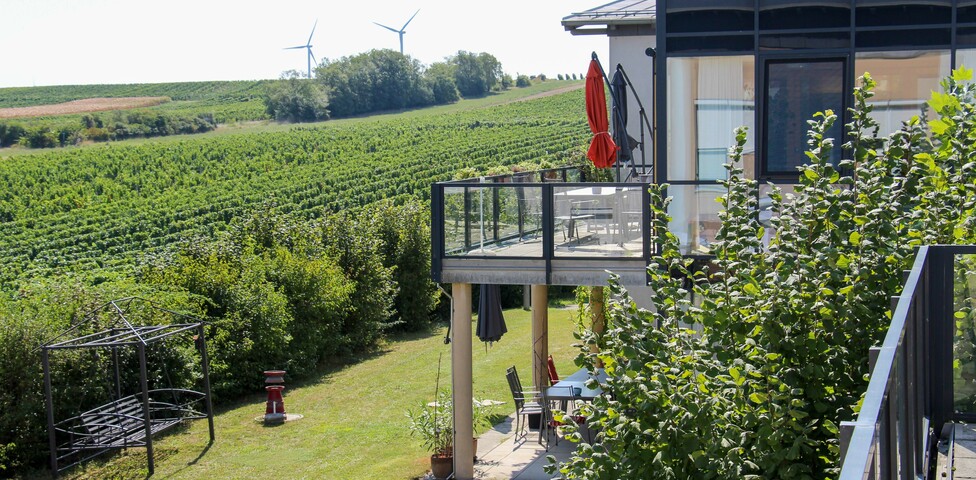 Das Bild zeigt den Balkon und Garten des Hauses Lisa mit Blick auf weitläufige Weinberge und Windräder in der Ferne. Die Umgebung wirkt grün und friedlich.