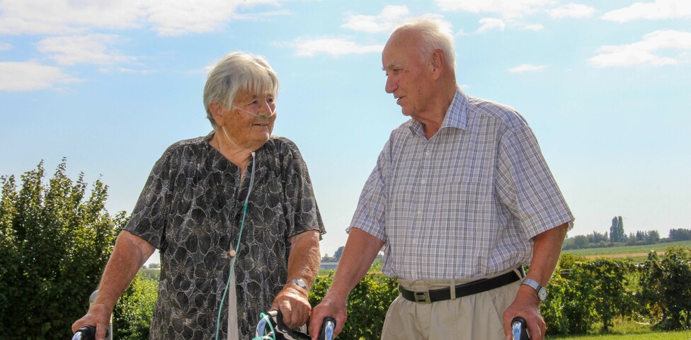 Zwei Personen aus dem Haus Lisa steht mit Rollatoren im Freien und unterhält sich. Die Frau hat eine Sauerstoffnasenbrille, und beide genießen den sonnigen Tag in einer ländlichen Umgebung.