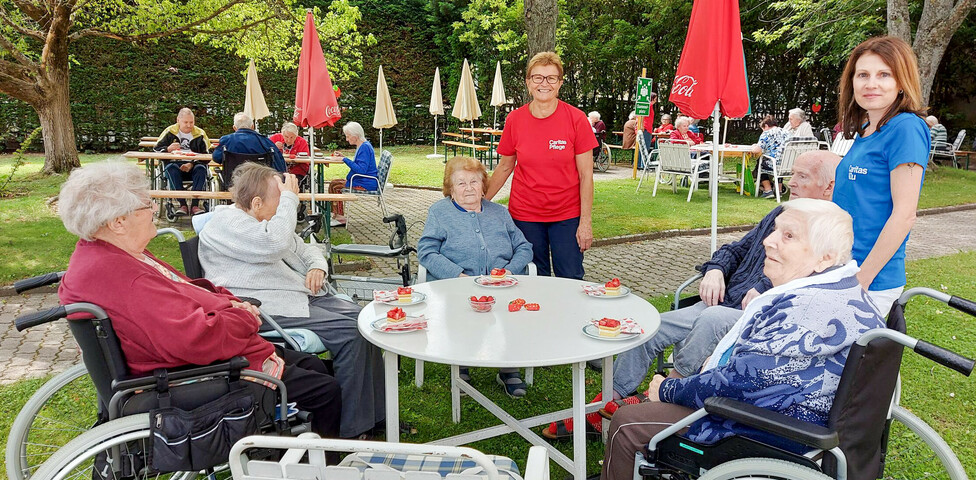 Man sieht die Bewohnerinnen des Haus Elisabeth mit Betreuerinnen. Sie genießen an einen Tisch im Freien das Erbeerfest. Es steht Erdbeerkuchen am Tisch