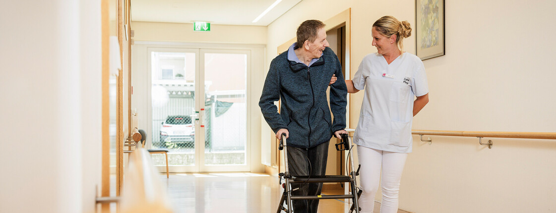 Eine Mitarbeiterin spaziert mit einem Bewohner am Rollator durch einen Gang unseres Pflegewohnhauses.