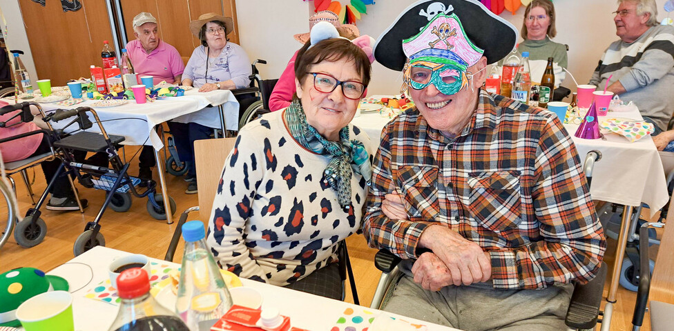 Ein Paar feiert fröhlich bei der Faschingsfeier im Haus Lisa. Der Mann trägt einen Piratenhut und eine bunte Maske, die Frau trägt weiße Katzenohren. Im Hintergrund sitzen weitere Gäste, umgeben von bunten Dekorationen.