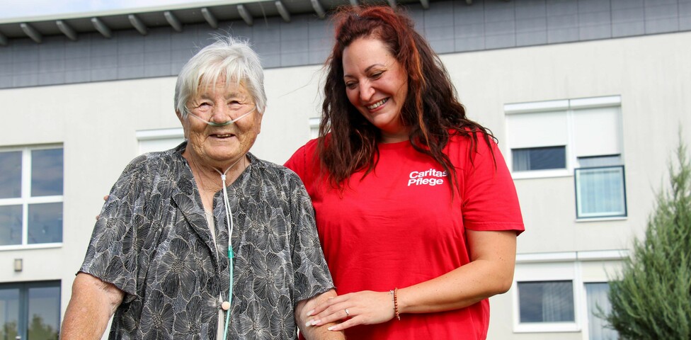 Eine Bewohnerin vom Haus Lisa, welche mit einem Gehwagen steht und Sauerstoffschläuche trägt. Neben ihr steht eine lächelnde Pflegekraft in einem roten T-Shirt mit der Aufschrift "Caritas Pflege", die sie unterstützend am Arm hält. Beide wirken zufrieden und fröhlich. 