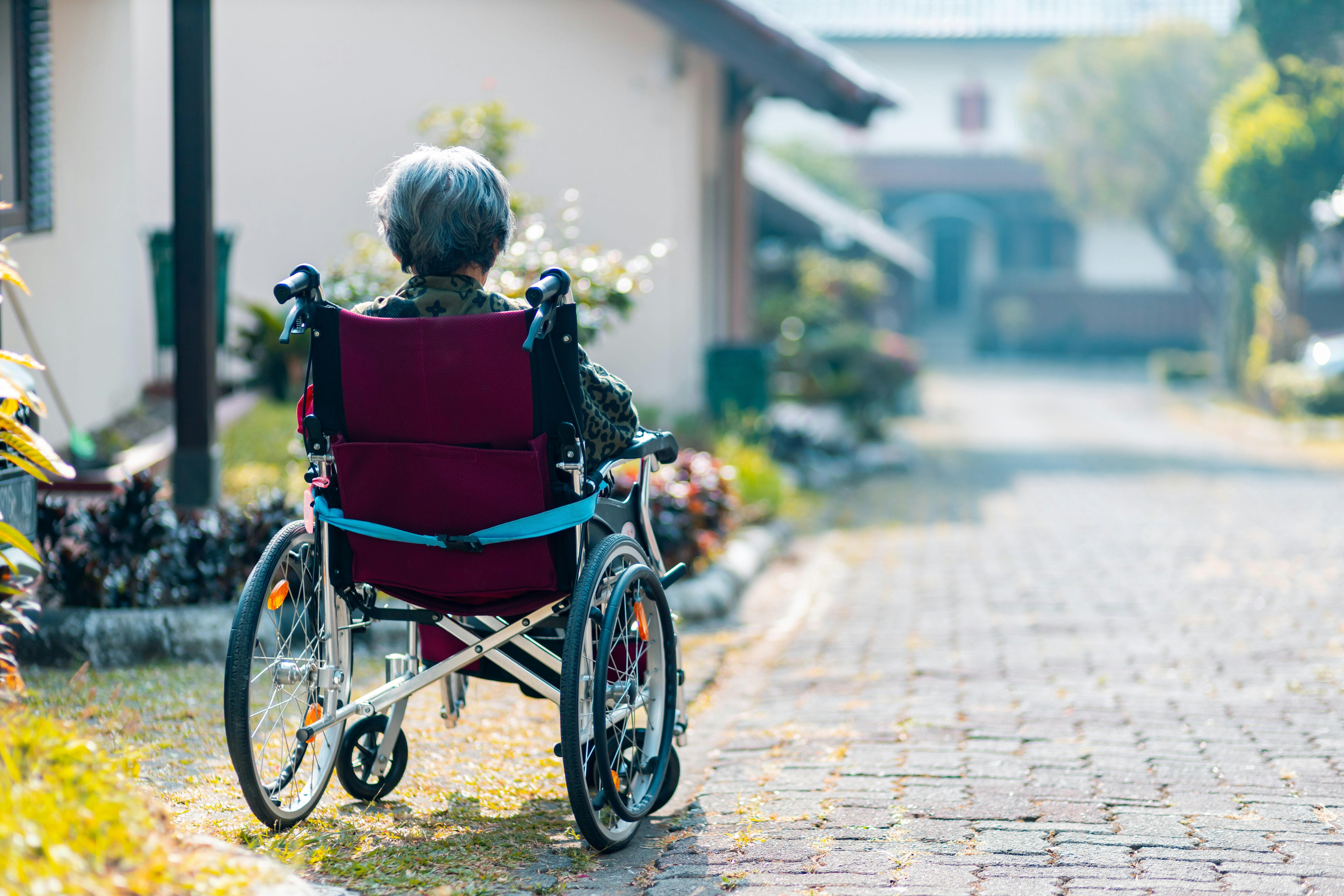 Eine im Rollstuhl sitzende Seniorin befindet sich in einer Hauseinfahrt und schaut die Straße hinunter.